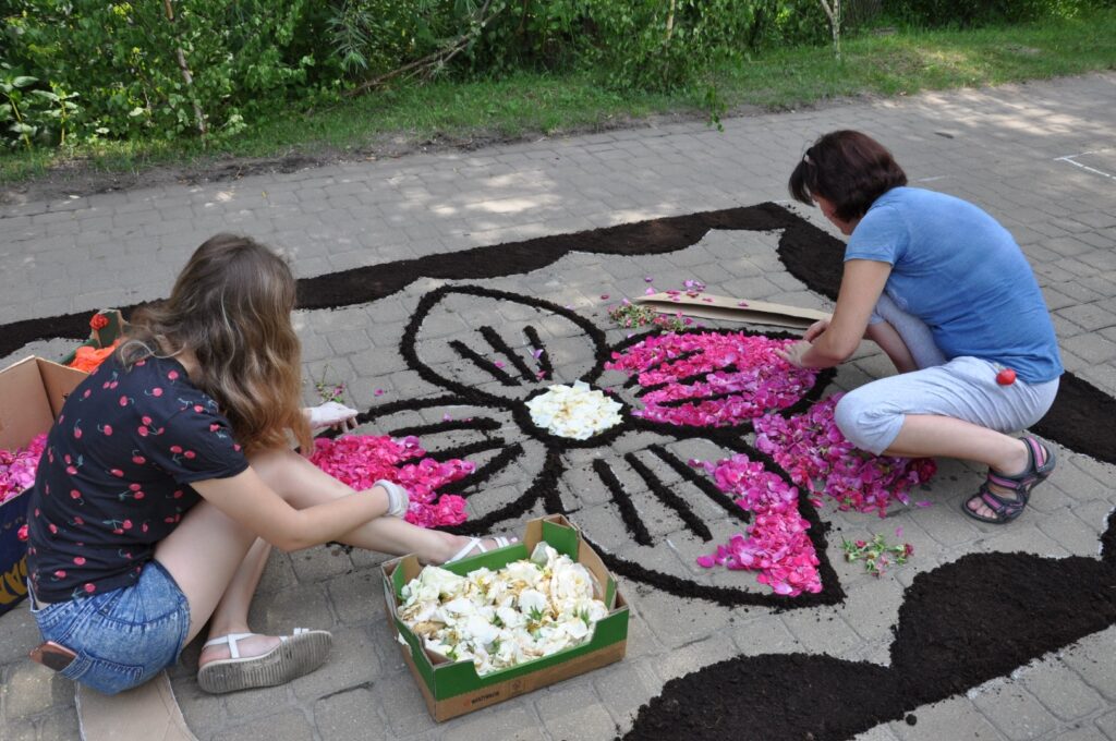 dwie dziewczynki układają fragment kwietnego dywanu. Usypują płtaki różowych kwiatów wewnatrz konturów.