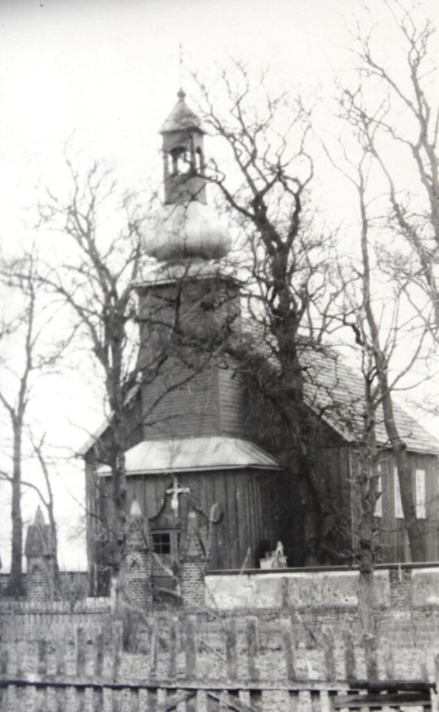 Drewniany kościół z wieżą zakończoną metalowym hełmem. Wokół kościoła drewniane ogrodzenie