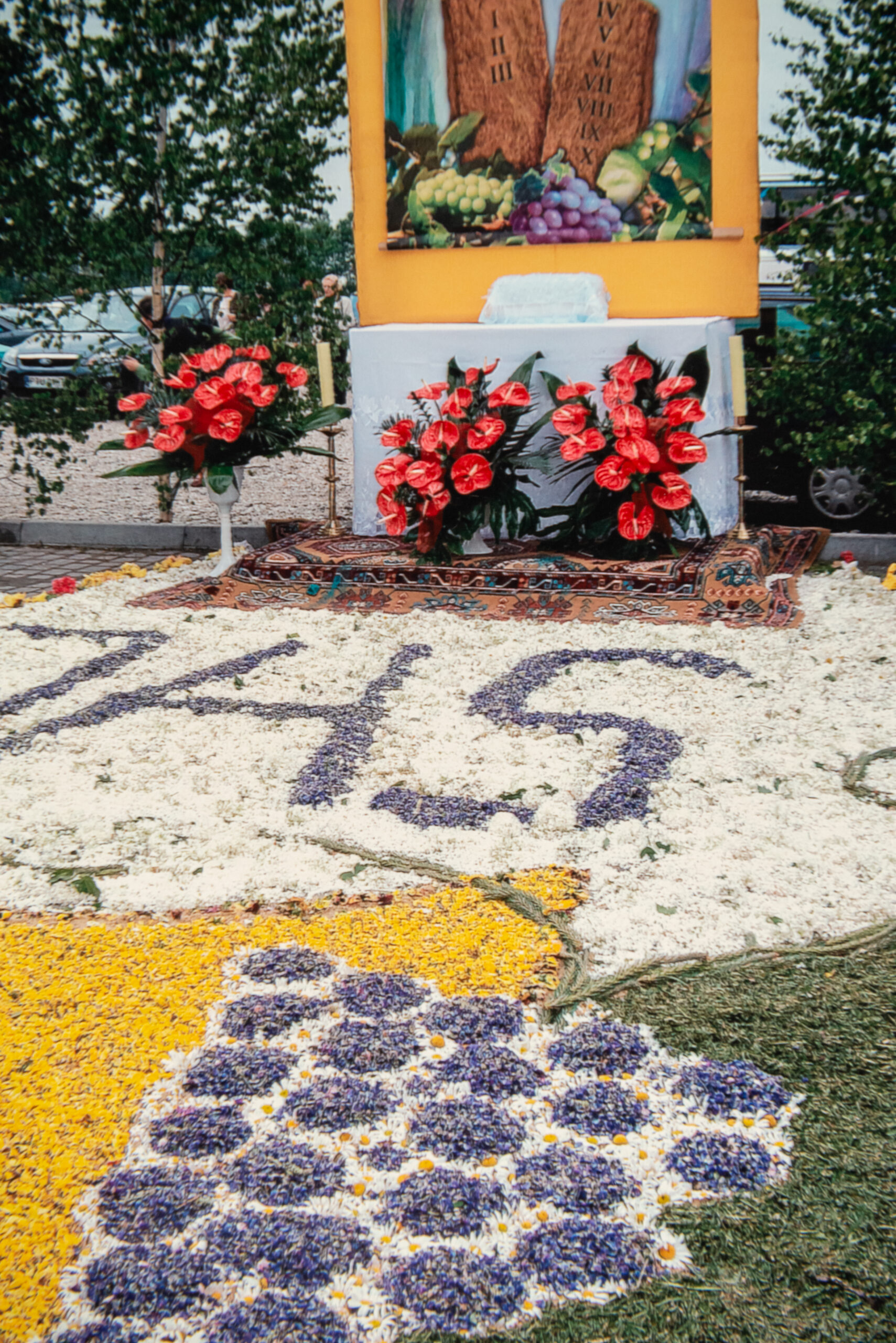 Kwiatowa kompozycja z białych, żółtych i fioletowych kwiatów o kształcie motywów winogron i hostii z napisem IHS. W tle ołtarz na trasie procesji ubrany przez mieszkańców Człop.