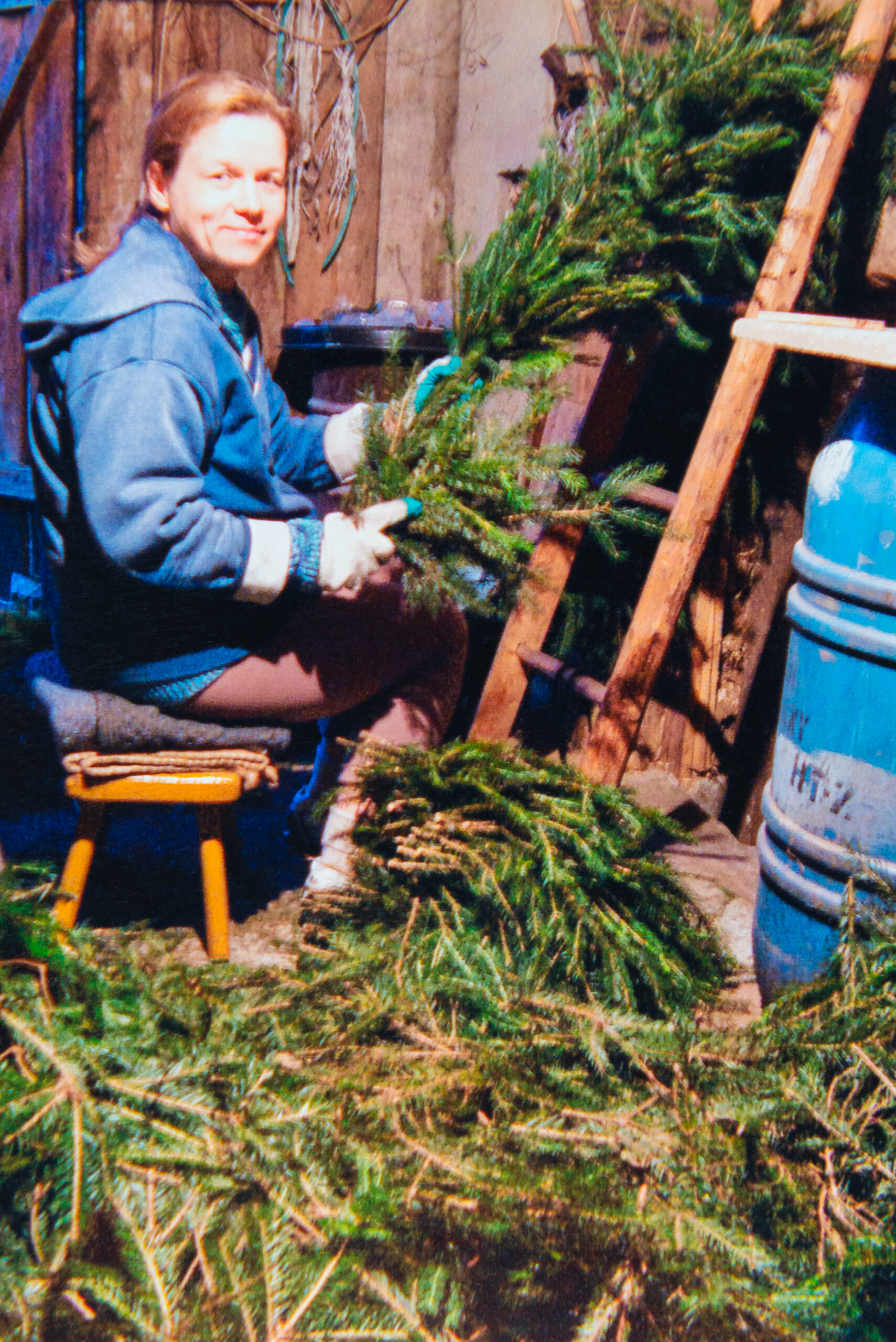 Kobieta w niebieskiej bluzie siedzi i przygotowuje gałęzie iglaste, duża ilość roślin spoczywa na ziemi.
