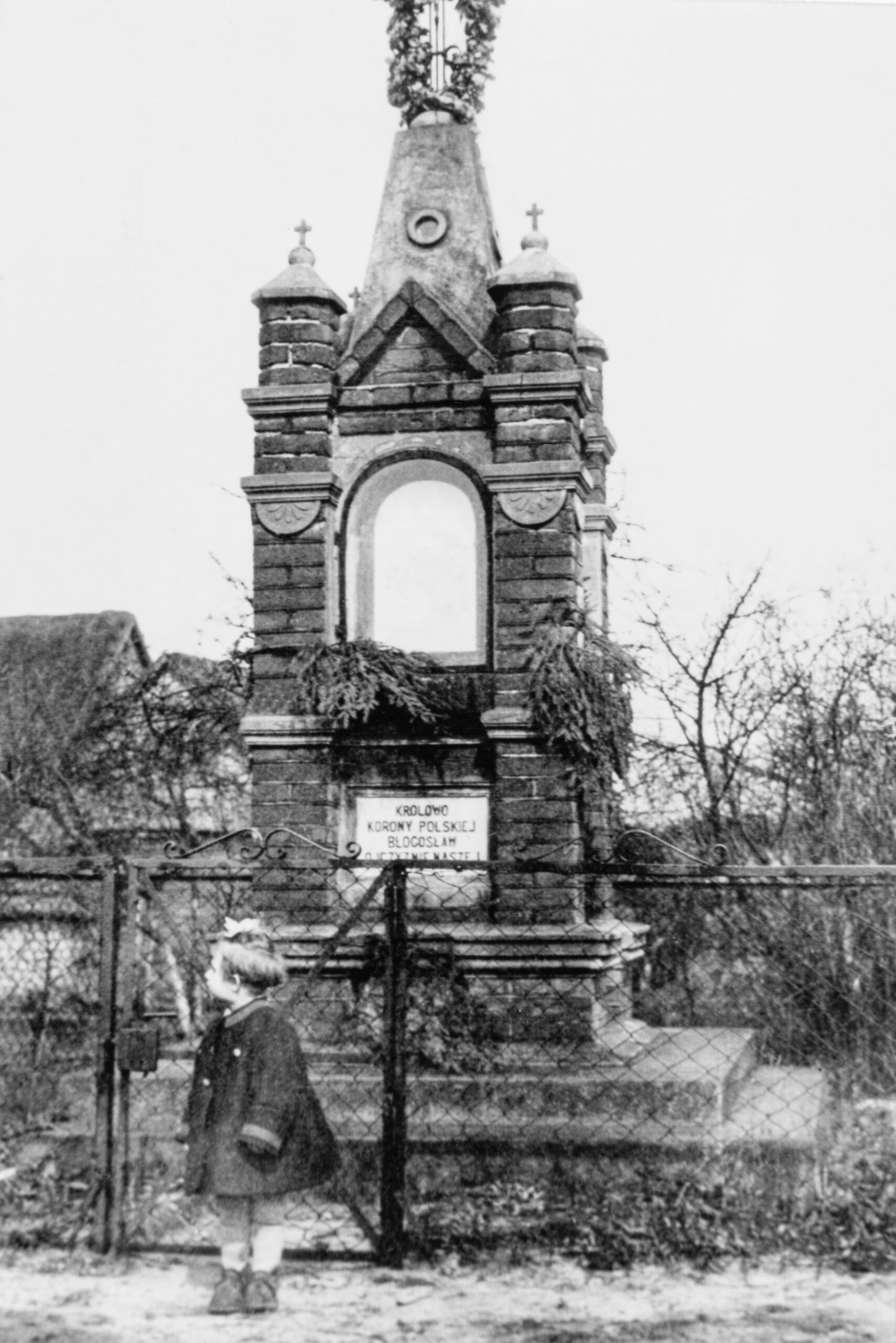 Czarno białe zdjęcie, dziewczynka pozująca na tle zabytkowej ceglanej kapliczki.