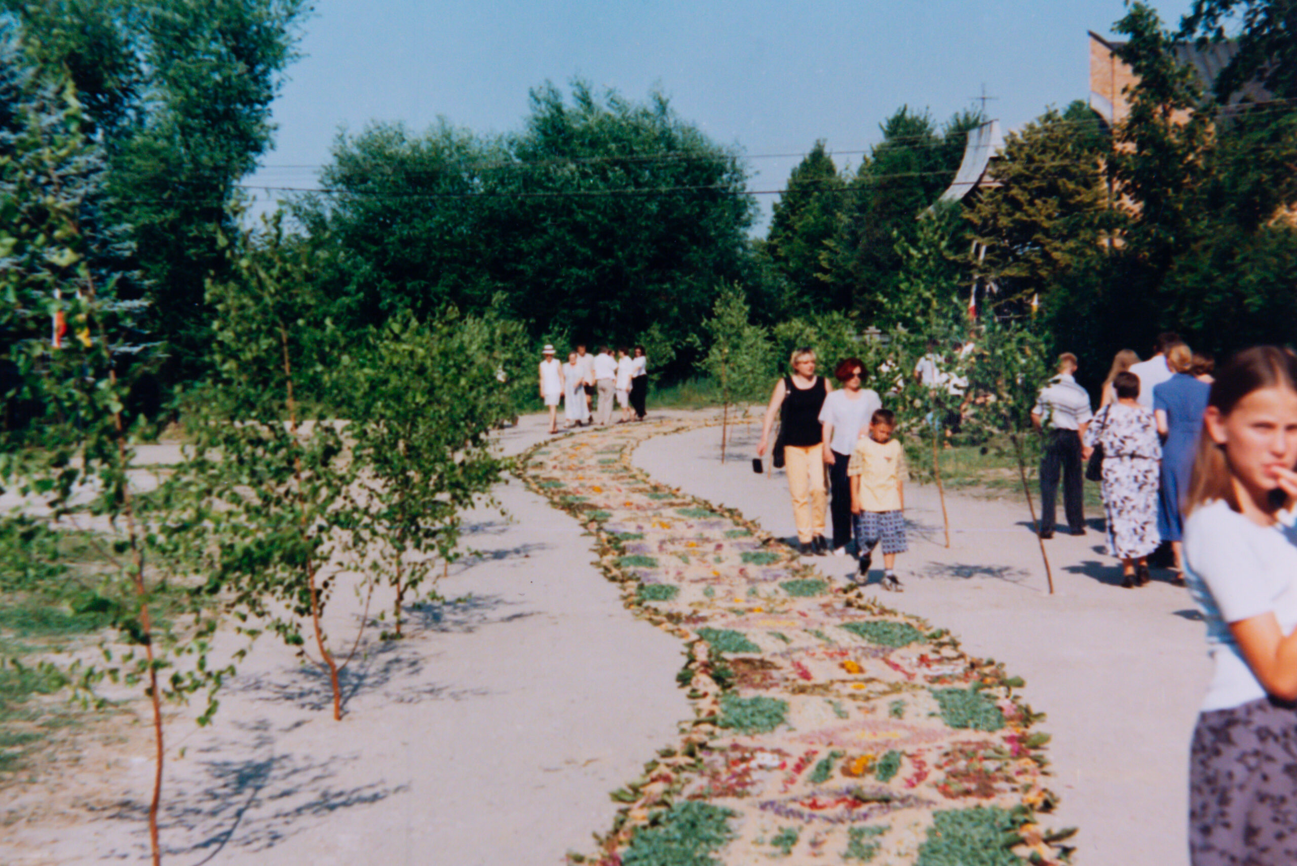 Kwietny dywan, złożony z wielokwiatowych kompozycji, obok drogi gałązki brzozowe wbite w piaszczysta drogę. Po prawej stronie oglądający datę ludzie.