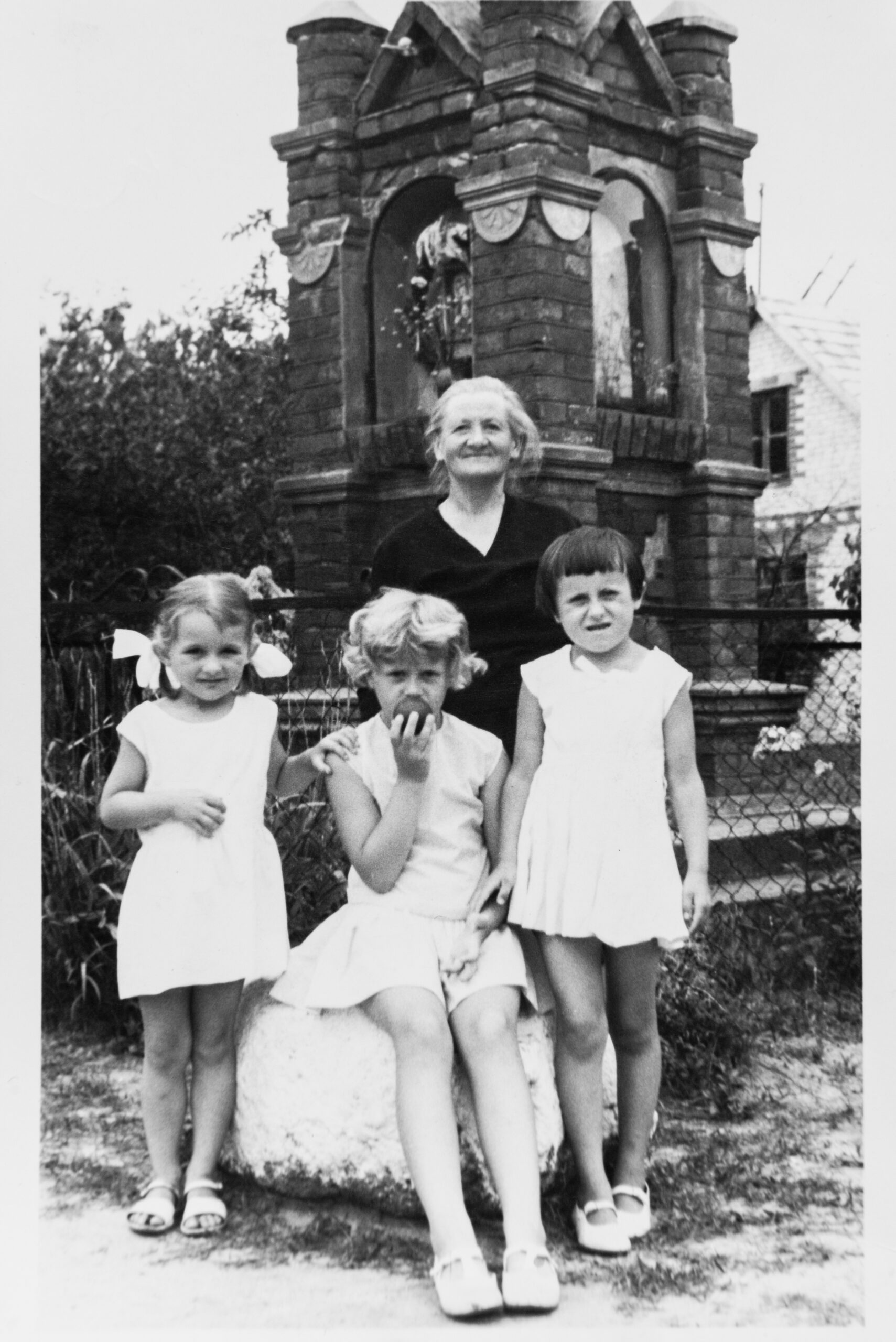 Zdjęcie czarno białe. Trzy dziewczynki oraz starsza kobieta pozują na tle starej ceglanej kapliczki.